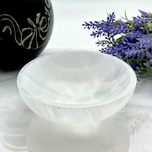 May include: A white selenite bowl, a type of crystal, is shown on a white surface. The bowl is smooth and has a slightly rounded shape.