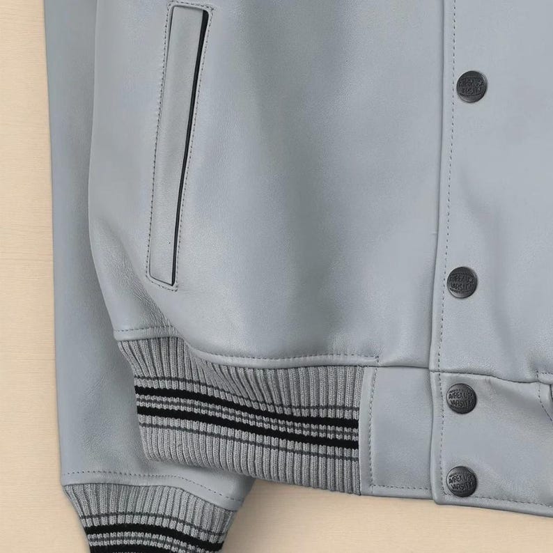 May include: A pale grey leather jacket with a ribbed knit hem and cuffs. The jacket has a pocket and black buttons. The cuffs have black stripes.