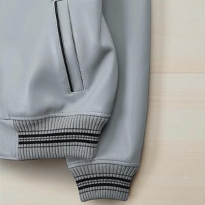 May include: A close-up of a pale grey leather jacket. The jacket has ribbed knit cuffs with two black stripes. A pocket with black trim is visible. The jacket is set against a light wood background.