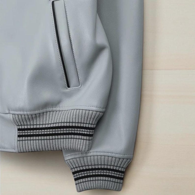 May include: A close-up of a pale grey leather jacket. The jacket has ribbed knit cuffs with two black stripes. A pocket with black trim is visible. The jacket is set against a light wood background.