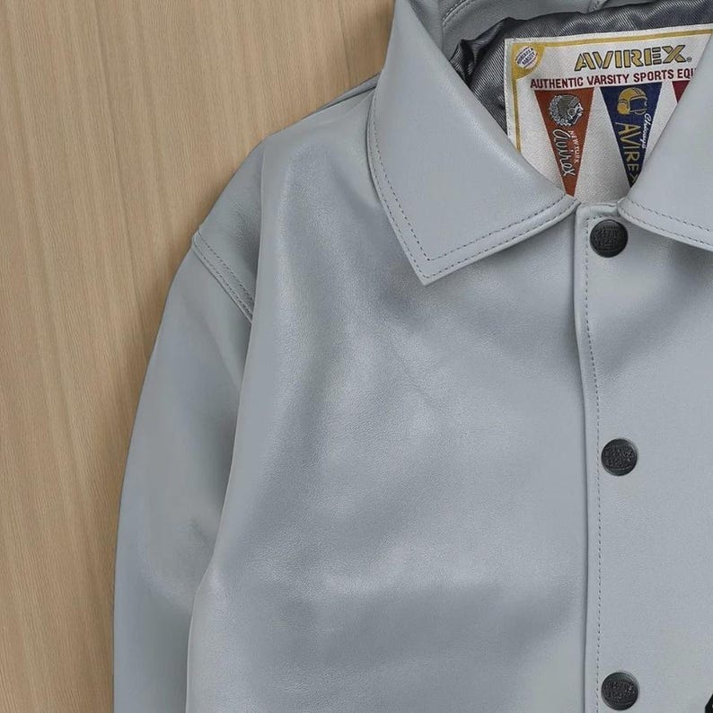 May include: A pale grey leather jacket with a traditional collar and black buttons. The jacket has a label with the text "AVIREX. AUTHENTIC VARSITY SPORTS EQ". The jacket is made of a smooth, matte material.