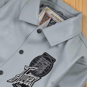 May include: A pale grey leather jacket with a button-down front and a collared neckline. The jacket has a large patch with the text "AVIREX" and "USA" on the front. A tag inside the collar reads "AVIREX AUTHENTIC VARSITY SPORTS".