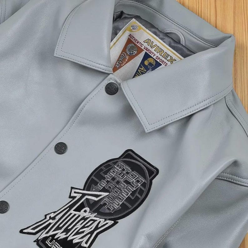 May include: A pale grey leather jacket with a button-down front and a collared neckline. The jacket has a large patch with the text "AVIREX" and "USA" on the front. A tag inside the collar reads "AVIREX AUTHENTIC VARSITY SPORTS".