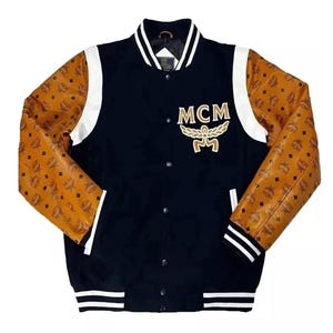 May include: A black varsity jacket with brown patterned sleeves and white accents. The jacket features the letters "MCM" in gold on the chest. The cuffs and hem have white stripes.