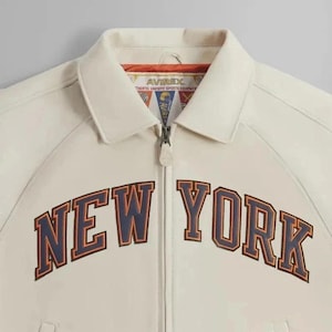 May include: Cream-colored jacket with the text "NEW YORK" in a bold, collegiate-style font across the chest. The jacket features a collared neckline, a full-length zipper, and the "AVIREX" logo near the collar.