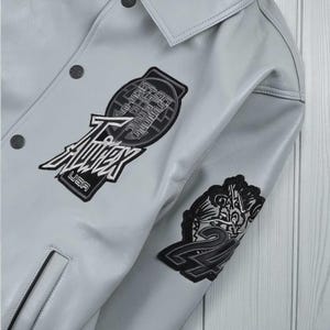 May include: A pale grey leather jacket with black embroidered patches. The jacket has a button-up front, a collar, and several patches with text and designs. One patch reads "T-Audex USA".