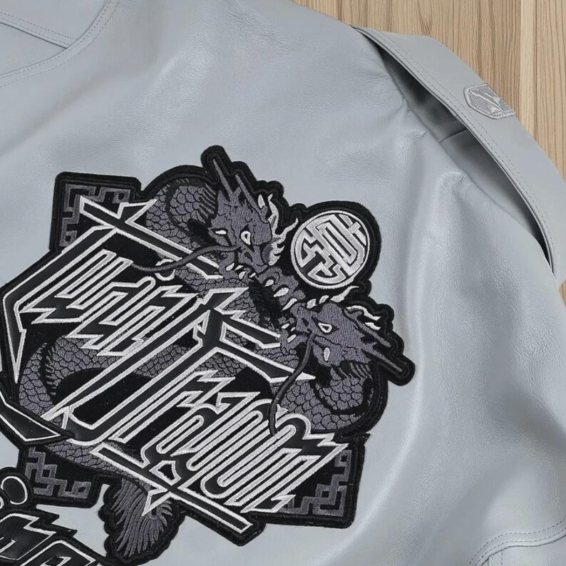 May include: A pale blue jacket with a large embroidered patch. The patch features two grey dragons and stylized text, outlined in black and white. A circular emblem is above the dragons.