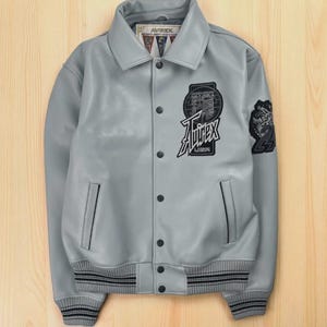 “Gray Avirex varsity jacket with front snap buttons, embroidered Avirex USA chest patch, sleeve patch detailing, ribbed cuffs and hem, and side pockets displayed on a wooden background.”