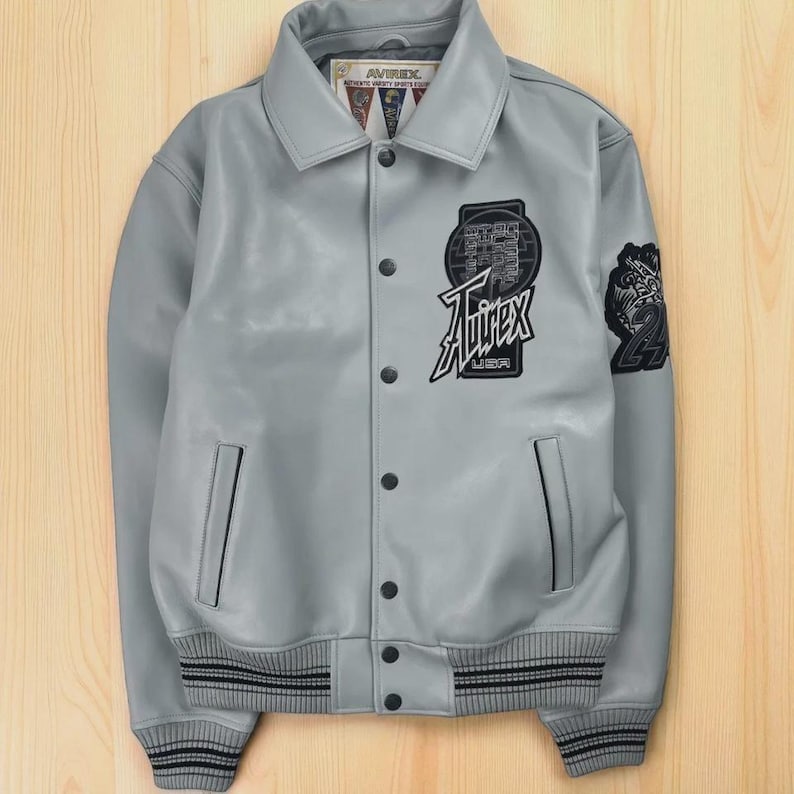 “Gray Avirex varsity jacket with front snap buttons, embroidered Avirex USA chest patch, sleeve patch detailing, ribbed cuffs and hem, and side pockets displayed on a wooden background.”