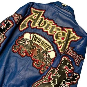 May include: A blue leather jacket with embroidered patches. The jacket features the brand name "Avirex" in large lettering, along with "New York" and lion graphics. The patches are outlined in red and gold.