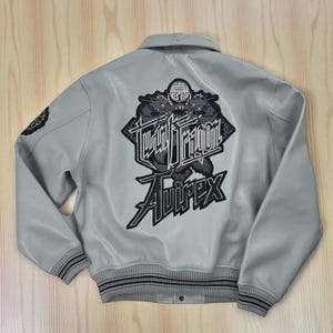 May include: Pale grey bomber jacket featuring a large black and white embroidered design of two dragons and the text "Twin Dragon" and "Avirex". The jacket has a ribbed collar, cuffs, and hem with black stripes.