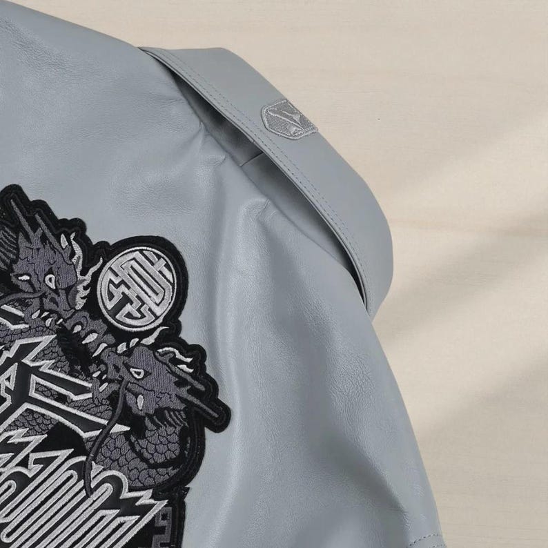 May include: A pale grey leather jacket with a detailed dragon embroidery in black, grey, and white. The jacket has a collar and a small emblem on the shoulder. The dragon design is on the front of the jacket.