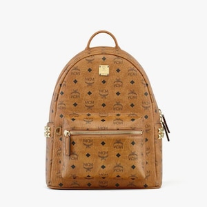 Può includere: Uno zaino marrone con un motivo ripetuto di diamanti neri e le lettere "MCM" e "WOW". Lo zaino ha una tasca frontale con cerniera dorata e borchie dorate sui lati. Una piccola placca dorata è visibile nella parte superiore.