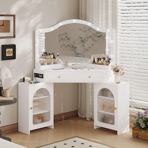 May include: A white vanity table with a large mirror framed by lights. The table has a drawer and two side cabinets with glass doors. Makeup and accessories are displayed on the tabletop. A vase of flowers sits on the side.