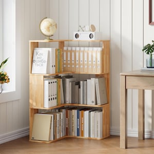 May include: A light-colored wooden corner bookshelf filled with books, binders, and decorative items. The bookshelf has three tiers and is positioned against a white wall. A globe and a small white speaker are on top.