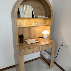 May include: A light oak wood desk with an arched hutch. The desk has two drawers with gold handles, a small white lamp, and a shelf with books. A water bottle and a notebook are on the desk. The desk is 60 inches tall.