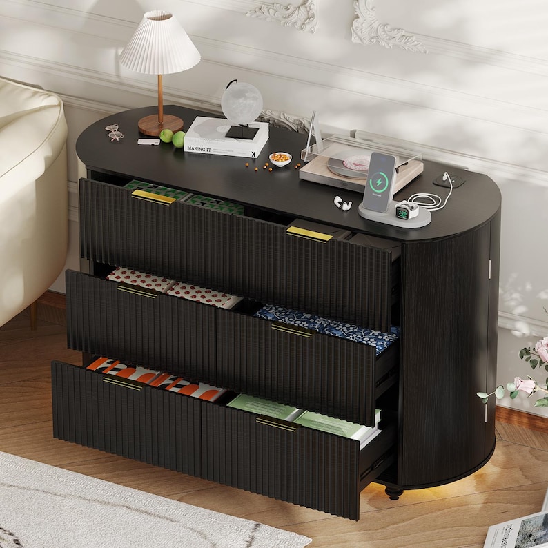 May include: A black, oval-shaped cabinet with three open drawers revealing colorful textiles. The cabinet has gold handles and a textured front. A lamp, globe, and record player sit on top.