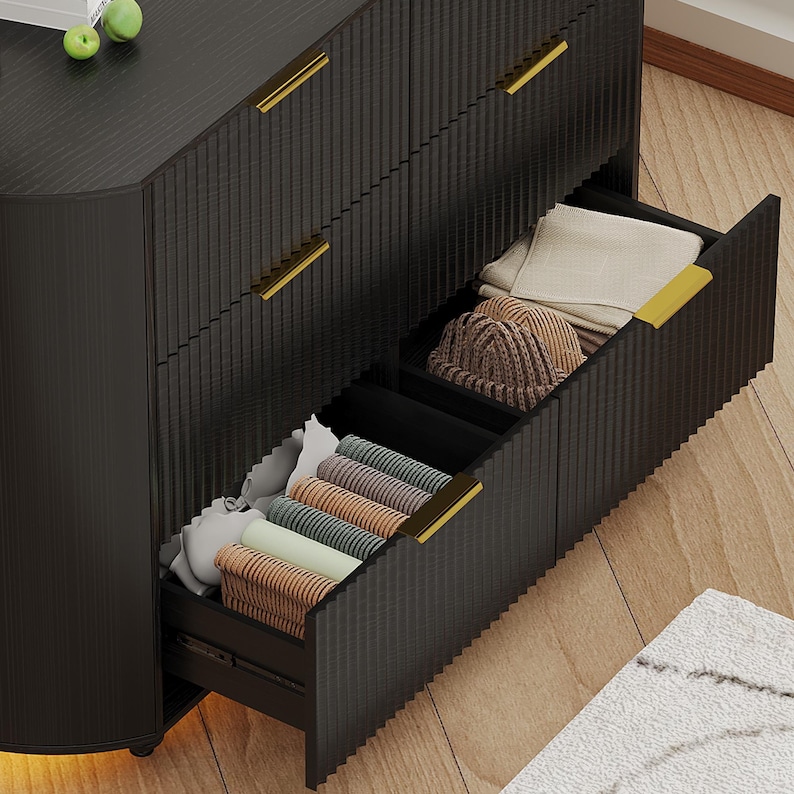 May include: A black dresser with gold handles. The open drawers reveal neatly folded clothing, including rolled-up towels and a knitted hat. The dresser has a textured, vertical-lined design.