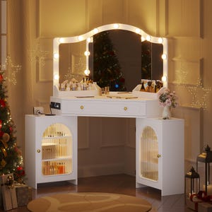 May include: A white vanity table with a large mirror framed by lights. The vanity has a drawer and two side cabinets with arched, clear doors. The table is decorated with makeup products and a vase of pink flowers. A Christmas tree is visible in the background.
