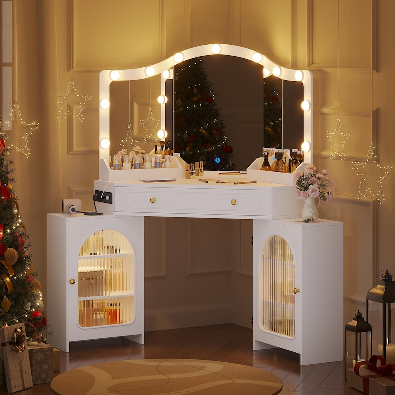 May include: A white vanity table with a large mirror framed by lights. The vanity has a drawer and two side cabinets with arched, clear doors. The table is decorated with makeup products and a vase of pink flowers. A Christmas tree is visible in the background.
