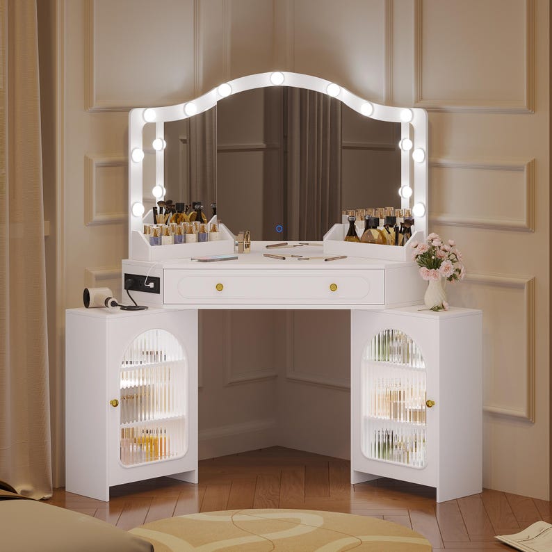 May include: White vanity table with a large, arched mirror framed by lights. The table has a drawer, two cabinets with glass doors, and gold-colored hardware. A vase of flowers sits on the right side.