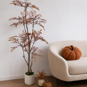 May include: A tall artificial tree with reddish-brown leaves in a textured white cylindrical pot, a cream-colored curved sofa with a round orange pumpkin-shaped pillow, and two small decorative pumpkins on the floor near the tree base.