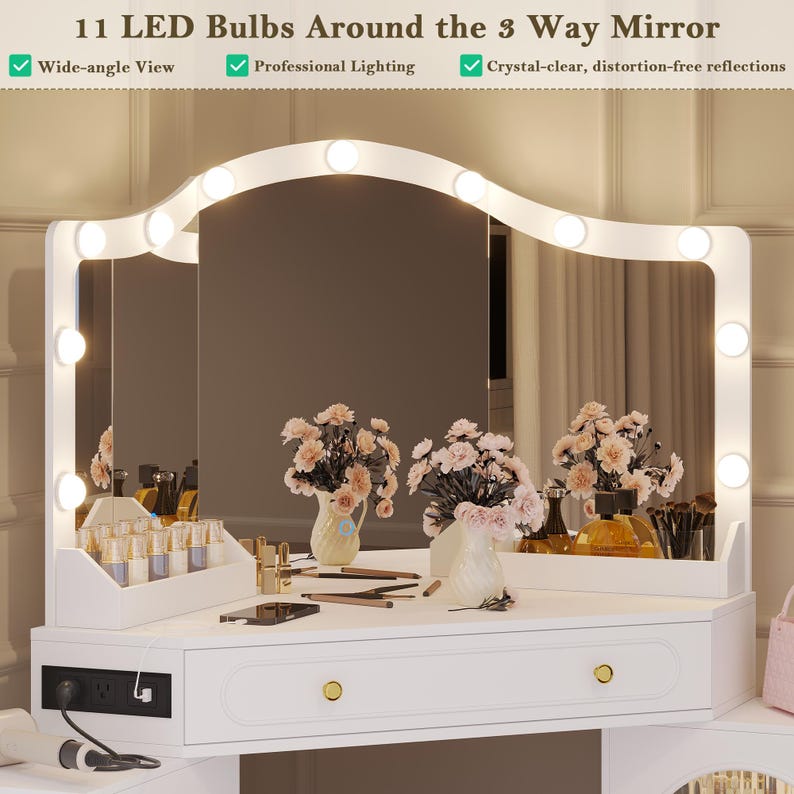 May include: A white vanity with a three-way mirror, illuminated by 11 LED bulbs. The vanity features drawers with gold knobs and is adorned with floral arrangements and beauty products. Text on the image reads: "11 LED Bulbs Around the 3 Way Mirror."