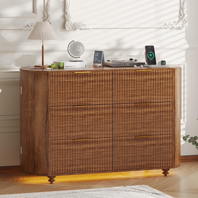 May include: A wooden dresser with six drawers and gold-colored handles. The dresser has a textured front and sits on short, decorative legs. A lamp, globe, and other items are on top.