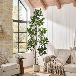 May include: A tall, artificial tree with vibrant green leaves stands in a white pot. The tree is placed in a bright, airy living room with a large window, a beige sofa, and a decorative throw blanket. The room has a neutral color palette.