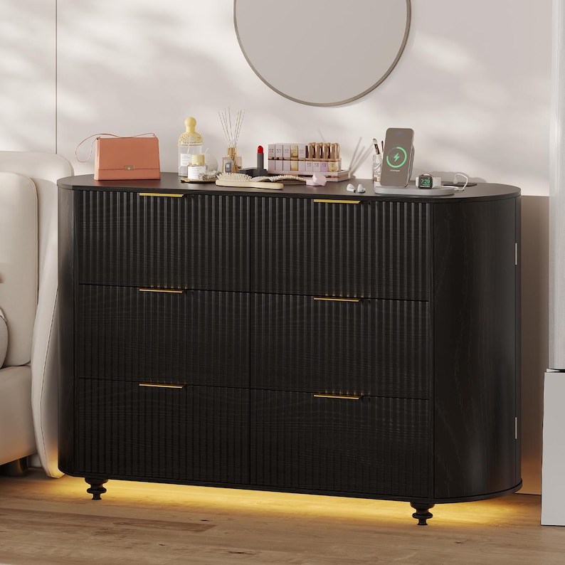 May include: A black six-drawer dresser with gold-toned hardware and a textured, vertical-grooved design. The dresser has a curved front and sits on short, black, decorative feet. A round mirror hangs above the dresser, which is topped with beauty products.