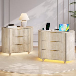 May include: Two light wood nightstands with three drawers each, gold handles, and a ribbed design. One has a lamp, the other a charging station with a phone and tablet. Both have a warm under-glow.