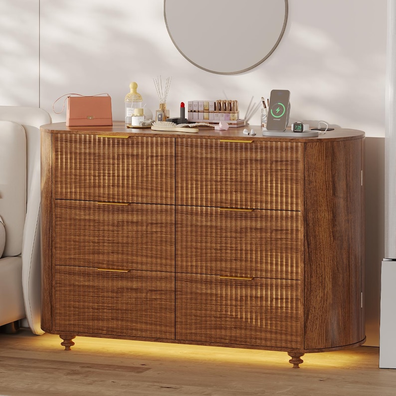 May include: A wooden dresser with six drawers and gold-colored handles. The dresser has a curved front and sits on small, decorative legs. The top of the dresser holds a mirror, cosmetics, and a charging phone. The dresser is illuminated with a warm light.