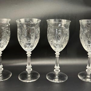 Puede incluir: Cuatro copas de vino transparentes con diseños florales grabados. Cada copa tiene un tallo largo y una base redonda. Las copas están dispuestas en fila sobre un fondo oscuro, mostrando su elegante diseño.