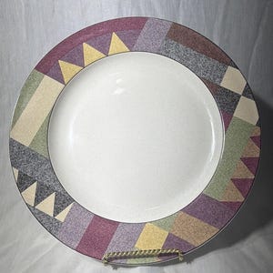 May include: A white ceramic dinner plate with a colorful geometric border. The border features triangles, squares, and rectangles in shades of purple, yellow, green, and gray. The plate is displayed on a gold-colored wire stand.