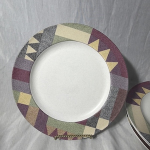 May include: A set of two white ceramic plates with a geometric border design. The border features a pattern of rectangles, triangles, and squares in shades of purple, green, yellow, and gray. The plates are displayed on a gold-colored metal stand.
