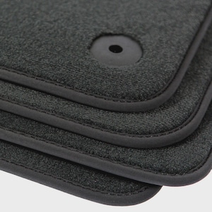 May include: Close-up of a stack of four dark gray car floor mats. Each mat has a textured surface, a black rubber border, and a circular fastener. The mats are designed to protect a car's interior.