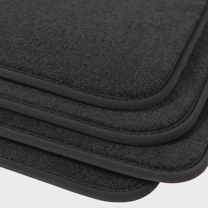 May include: Close-up of a stack of four black floor mats. Each mat has a textured surface and a rounded corner with a black border. The mats are made of a soft, absorbent material.