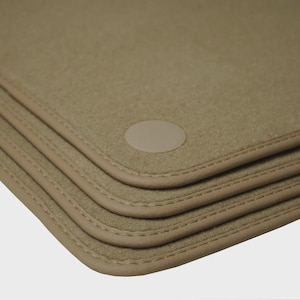May include: Tan car floor mats with a stitched border and a circular patch. The mats are stacked, showing the layered design. The color is a neutral beige.