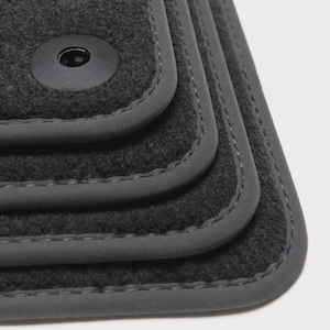 May include: Close-up of a set of dark gray car floor mats. The mats have a textured, felt-like surface with a smooth, gray border and stitching. A black circular fastener is visible on the top mat.