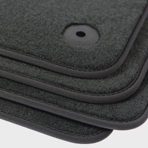 May include: Close-up of a stack of four dark gray car floor mats. Each mat has a textured surface, a black rubber border, and a circular hole with a black grommet. The mats are designed to protect a car's interior.