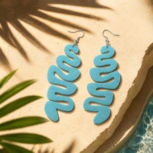 May include: A pair of dangling earrings featuring a long, light blue, wavy, serpentine shape with a textured surface. Each earring is attached to a silver-toned metal hook, creating a fluid, elongated design that hangs vertically from the ear wire.