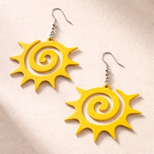 May include: A pair of yellow sun-shaped earrings. Each earring features a spiral center and pointed rays, suspended from silver-colored fish hook ear wires. The earrings are displayed against a light background.