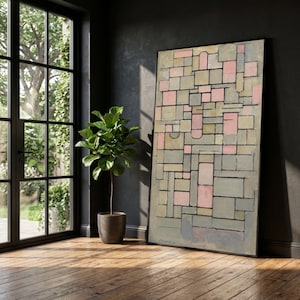 Könnte beinhalten: Abstraktes Gemälde mit schwarzem Rahmen, das ein geometrisches Design aus rosa, grauen und beigen Rechtecken und Quadraten zeigt. Das Kunstwerk lehnt an einer dunkelgrauen Wand, neben einer Topfpflanze und einem Fenster.