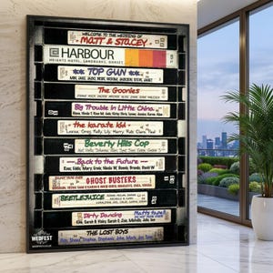 Puede incluir: Una obra de arte enmarcada que presenta una colección de cintas VHS vintage, cada una etiquetada con un título de película como "Top Gun" y "Regreso al futuro". Las cintas están dispuestas verticalmente, con el texto "WEDFEST" en la parte inferior.