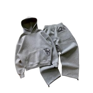 Puede incluir: Un conjunto de sudadera con capucha y pantal&oacute;n de ch&aacute;ndal grises. La sudadera tiene un bolsillo delantero y una capucha. El pantal&oacute;n de ch&aacute;ndal tiene un bolsillo en la parte trasera. Ambas piezas tienen un peque&ntilde;o dise&ntilde;o bordado. El conjunto es de material suave y c&oacute;modo.