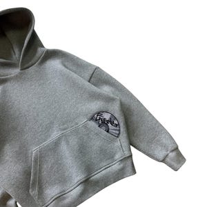 Puede incluir: Una sudadera con capucha gris con bolsillo de canguro y capucha. La manga izquierda tiene un parche circular con la palabra "Episodes" y un dise&ntilde;o de CD. La sudadera est&aacute; hecha de un material suave.