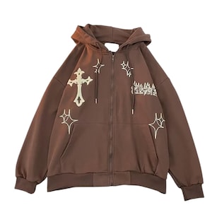 May include: A brown zip-up hoodie with a hood and front pockets. The hoodie features white embroidered crosses and starburst designs. The text on the right side of the chest is partially visible. The hoodie is made of a soft material.
