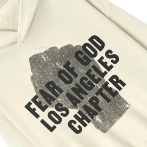 May include: Cream-colored hoodie with the text "FEAR OF GOD LOS ANGELES CHAPTER" in a distressed black font. The text is printed over a textured, gray rectangular shape. The hoodie has a hood and a relaxed fit.