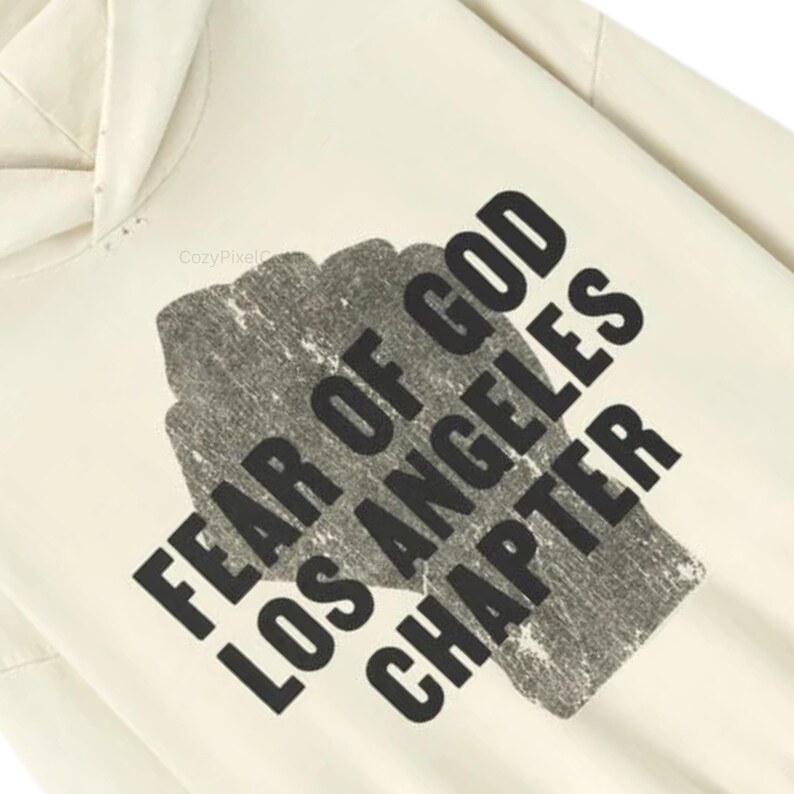 May include: Cream-colored hoodie with the text "FEAR OF GOD LOS ANGELES CHAPTER" in a distressed black font. The text is printed over a textured, gray rectangular shape. The hoodie has a hood and a relaxed fit.
