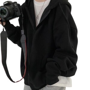 May include: A black zip-up hoodie with a hood and front pockets. The hoodie is worn over a white t-shirt. A black camera with a strap is held in the left hand.